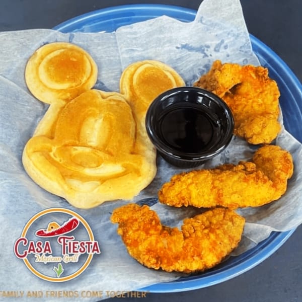 Mickey Mouse shaped waffle with chicken tenders and a side of syrup on a blue plate.