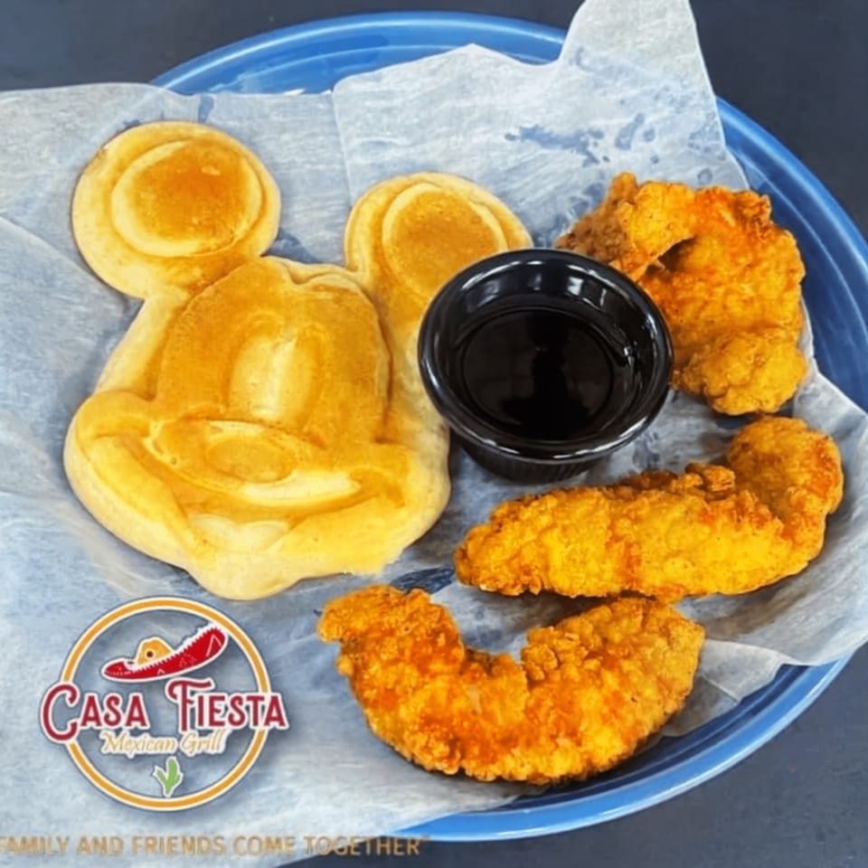 Mickey Mouse shaped waffle with chicken tenders and a side of syrup on a blue plate.